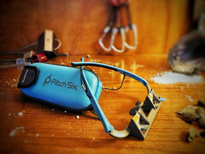 EyeSend belay glasses with case on a wooden background. Quickdraws, nuts and climbing chalk in the background of the belay glasses.